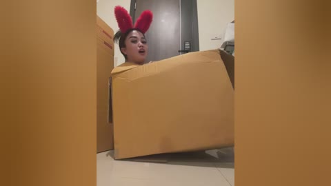 Media: Video of a young Asian woman with short black hair wearing a yellow jumpsuit and red bunny ears, hiding in a cardboard box on a tiled floor, with a black fridge and white toilet visible in the background.