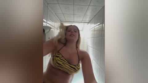 Media: Video of a blonde woman in a yellow and black striped bikini, showering in a white-tiled bathroom with a tiled ceiling.