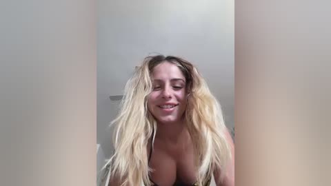 Media: Video of a smiling, topless, light-skinned woman with long, blonde hair, partially obscured by vertical, beige curtains.