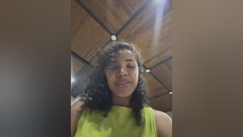 Media: Video of a young African-American woman with curly hair, wearing a bright green tank top, smiling indoors under a wooden ceiling with a lens flare effect.