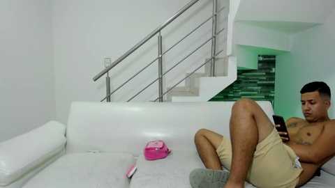 Media: Video of a shirtless, light-skinned man with short hair, wearing beige shorts, sitting on a white leather couch, texting on a phone. Background shows white walls, stairs, and a pink toy on the couch.