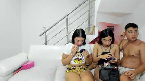 Media: Video of three young people, two women and a man, sitting on a white couch, playing video games. The background shows a modern, white stairway and minimalist decor.