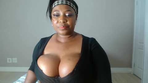 Media: Video of a dark-skinned Black woman with large breasts, wearing a black top that exposes them, in a plain room with beige walls and a white door.