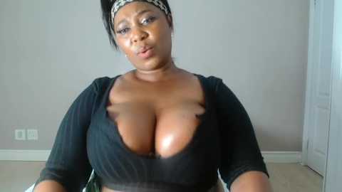 Media: Video of a plus-sized Black woman with a medium brown complexion, wearing a low-cut black top that accentuates her large breasts. She has a headband and is indoors, in a plain room with white walls and a door.