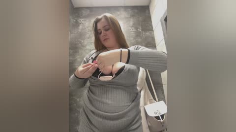 Media: A video of a curvy, fair-skinned woman with shoulder-length blonde hair in a gray sweater, revealing a large, black bra. She is adjusting the bra in a narrow, tiled restroom.