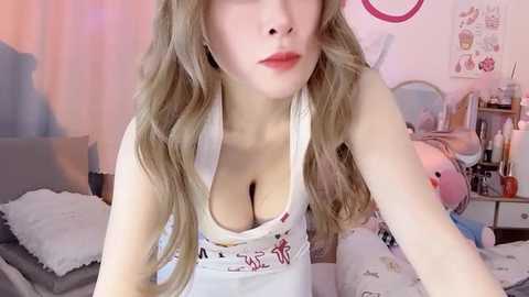 Video of a fair-skinned woman with long, wavy blonde hair, wearing a low-cut white tank top revealing ample cleavage, in a pink-themed bedroom with plush toys and posters.