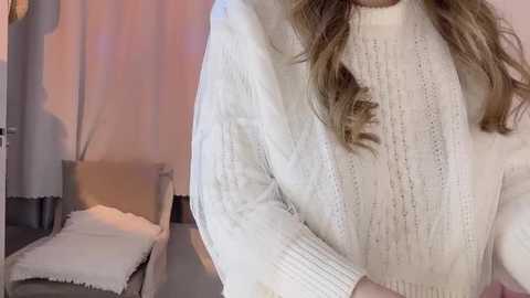 Media: Video of a woman in a cozy, white, knitted sweater with a high neck, seated on a light-colored, cushioned chair with a white blanket, in a softly lit, minimalist room.