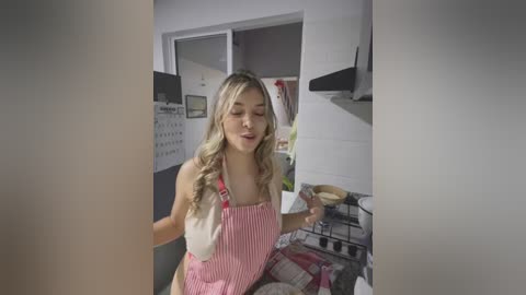 Media: A video of a blonde woman with long, wavy hair, wearing a pink striped apron, dancing in a kitchen with white walls and modern appliances.