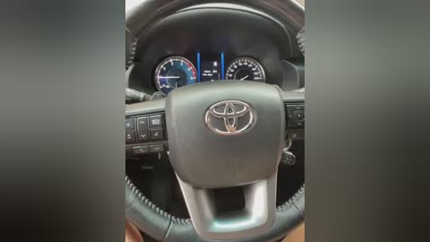 Media: Video of a Toyota SUV dashboard, featuring a black leather steering wheel with the Toyota emblem, digital speedometer, and gear shift on the left. The background displays a blurred interior with a black steering column and gear selector.