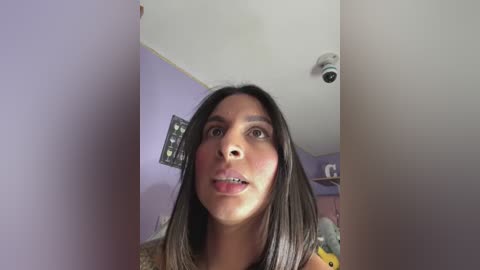 A close-up video of a young woman with long, dark hair and light skin, wearing a beige top, in a room with lavender walls and a ceiling light.