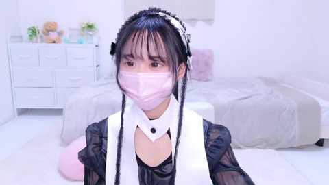 Media: Video of an Asian woman in a maid outfit, white mask, and black headband, kneeling on a white floor in a bright, tidy bedroom with a white dresser and plush toys.