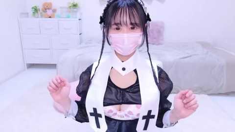 Media: Video of an Asian woman in a revealing black maid outfit with white cross straps, wearing a pink face mask, and a white headband with bunny ears. Background shows a white dresser with teddy bears and a bed.