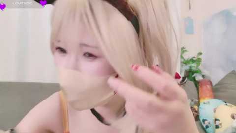 Media: A close-up video of a young woman with blonde hair, wearing a yellow mask, red nail polish, and a black choker, in a cozy room with plants and a soft bed.