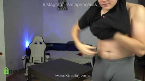 Media: Video of a plus-sized, light-skinned woman in a black crop top and gray pants, lifting her top, revealing a bare, round belly. Background includes a gaming chair, a table, and a bed. Text overlay reads: \"Instagram @sofia_princesss.\