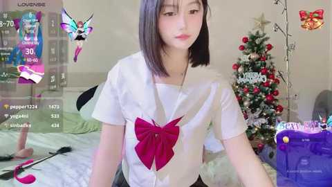 Media: Video of an Asian woman with straight black hair, wearing a white blouse with a red bow, sitting in a bedroom with a decorated Christmas tree.