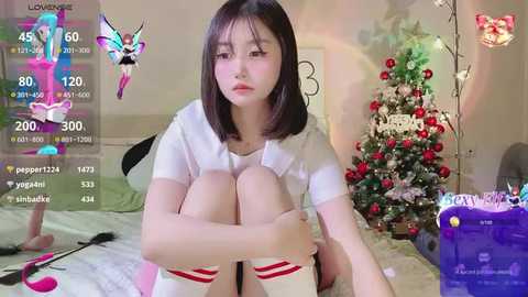 Media: Video of an East Asian woman with shoulder-length black hair, wearing a white t-shirt and knee-high socks, sitting on a bed with a festive Christmas tree in the background.