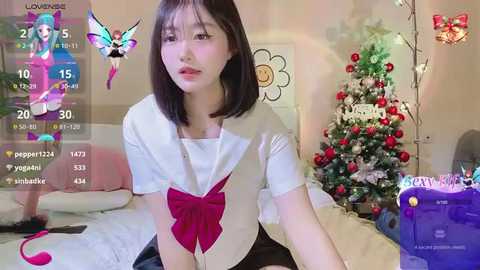 Media: Video of an Asian woman with straight black hair, wearing a white blouse with a red bow, sitting in a cozy room with a Christmas tree and digital game interface.