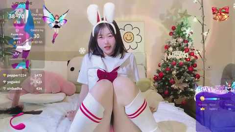 Media: A young East Asian woman with straight black hair, wearing a white bunny costume, sits on a bed with a Christmas tree in the background. The image has a playful, festive atmosphere.