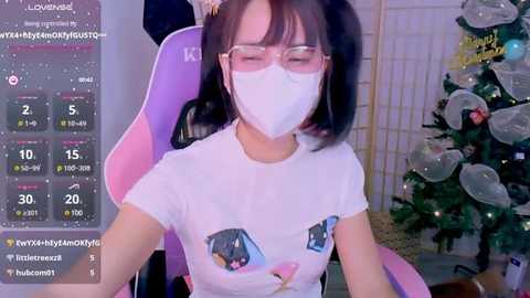 Media: Video of an Asian woman with pigtails, wearing a white T-shirt with cartoon characters, a face mask, and glasses, sitting on a pink gaming chair in a cozy room with a decorated Christmas tree.