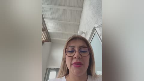 Media: A video of a young woman with long, straight, blonde hair, wearing rectangular glasses and a white shirt. She stands indoors, with a white ceiling and exposed ductwork in the background.