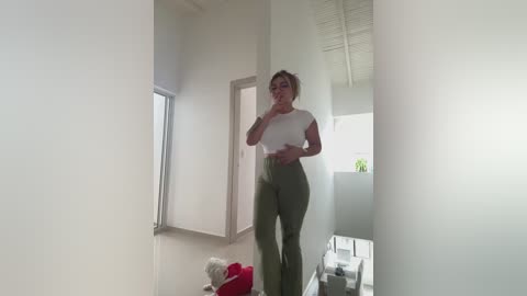 Media: Video of a woman with glasses, wearing a white crop top and olive-green pants, taking a selfie in a minimalist, brightly lit room.