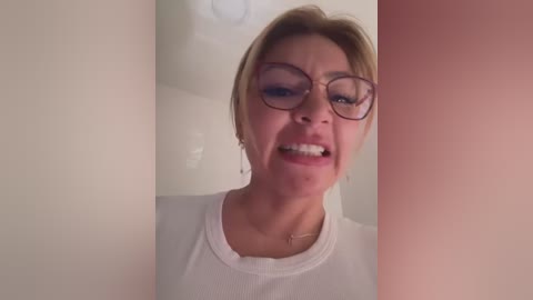 Media: Video of a blonde woman with short hair, wearing glasses and a white tank top, smiling with her mouth open, in a dimly lit room with cream-colored walls.