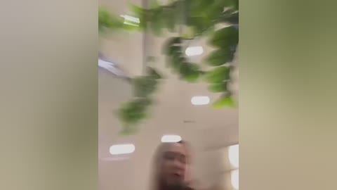 Media: A blurry video showing a young woman with light skin and long blonde hair, standing in a brightly lit indoor space with green hanging plants.