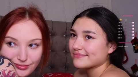 Media: Video of two smiling young women with fair skin and red hair (left) and black hair (right), in a cozy room with a gray tufted headboard.