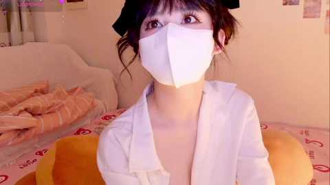 Media: Video of an Asian woman with fair skin, dark hair, and large eyes, wearing a white face mask, open white shirt, and a black headband, sitting on a bed with a plush toy, in a cozy, dimly lit room.