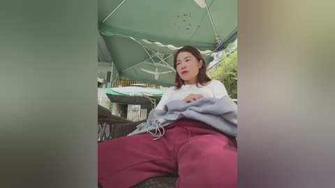 Media: Video of an East Asian woman with short brown hair, wearing a white top, gray sweatshirt, and pink sweatpants, sitting under a green umbrella, in a park setting.