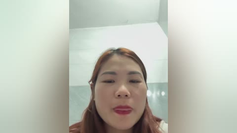 Media: Video of a young Asian woman with straight, shoulder-length reddish-brown hair, light skin, and subtle makeup. She is indoors, facing the camera with a neutral expression. The background is a minimalist, white-walled room.