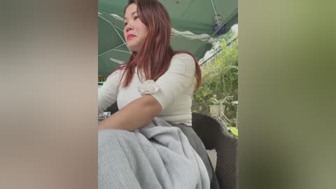 Media: Video of an Asian woman with medium-length auburn hair, wearing a white, long-sleeved top and a light gray sweater, seated outdoors under a green umbrella, with lush greenery in the background.