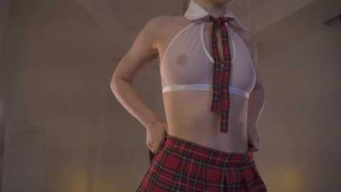 Media: Video of a light-skinned woman wearing a revealing, white halter crop top and red plaid skirt, standing in a dimly lit, minimalist room.