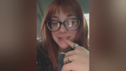 Media: Video of a young woman with fair skin and long, straight red hair, wearing black-framed glasses, biting her index finger, and holding a green ring. Background is blurred.