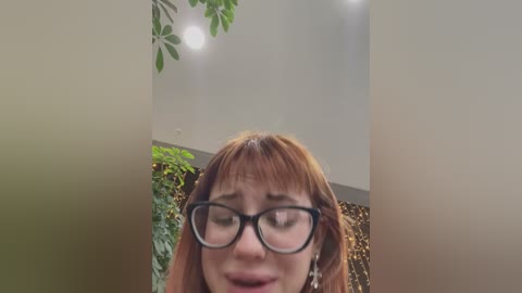 Media: Video of a close-up, blurred face with glasses, red hair, and a worried expression, framed by greenery and a beige wall, capturing a candid moment.