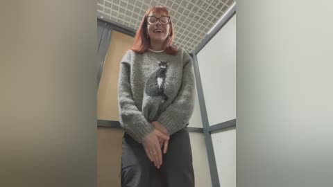 Media: Video of a woman with red hair, glasses, wearing a grey sweater with a cat design, and black pants, standing in a small, dimly lit room with white ceiling tiles.