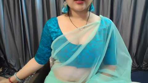 Media: Video of a fair-skinned, middle-aged woman wearing a blue saree with a turquoise blouse, teal earrings, and a black necklace. She sits against gray drapes, with a subtle smile.