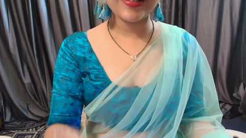 Media: Video of a woman wearing a vibrant blue, semi-transparent sari with a floral-patterned blouse. She has a light complexion, red lipstick, and a delicate necklace. The background features gray, flowing curtains.