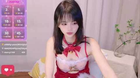 Media: Video of an Asian woman with long black hair, wearing a revealing Santa outfit, sitting on a bed with a white sheet, against a plain background.