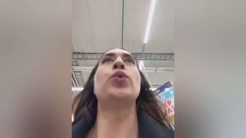 A video of a woman with light skin, long dark hair, and a black blazer, making a kissy face towards the camera, taken in a bright, modern room with fluorescent lights and a ceiling grid.