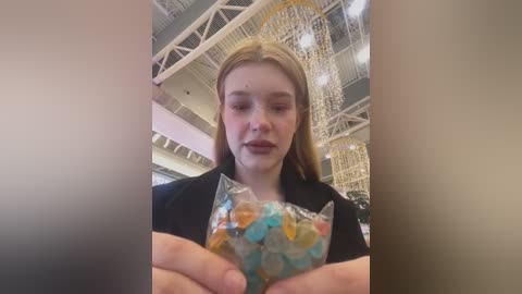 Media: Video of a young woman with light skin and long blonde hair, wearing a black jacket, holding a clear plastic bag filled with colorful candy. Background shows a large, ornate chandelier and a high-ceilinged indoor space.