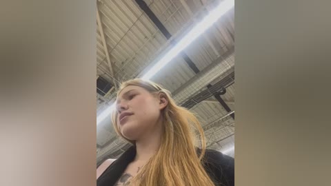 Media: Video of a young Caucasian woman with long, straight blonde hair, wearing a black top, standing in a dimly lit, industrial warehouse with exposed beams and fluorescent lighting.