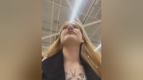 Media: Video of a young woman with long blonde hair, wearing a black blazer, standing in an industrial setting with a white ceiling and metallic beams.