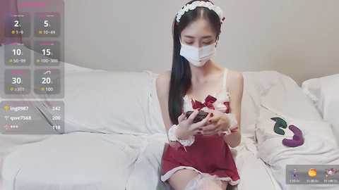 Media: A video of an Asian woman in a red maid outfit, white face mask, and white headband, sitting on a bed with white sheets, wearing a VR headset, holding a smartphone.