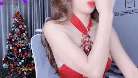 Media: Video of a woman with fair skin, long brown hair, and red lipstick, wearing a red halter dress, sitting in a room with a decorated Christmas tree and festive wall art.