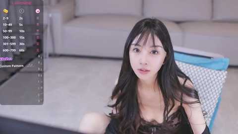 Media: A video of an East Asian woman with long black hair and fair skin, wearing a black lace bra, sitting on a cushion in a modern living room.