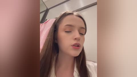 Media: Video of a young Caucasian woman with straight brown hair, closed eyes, and a slight smile. She is wearing a white shirt and appears to be indoors, possibly in a bathroom.