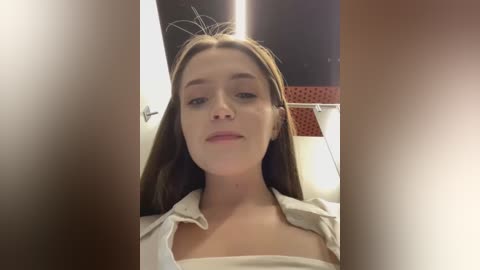 Media: Video of a young Caucasian woman with light skin and straight brown hair, wearing a white shirt, seated indoors with a dimly lit background.