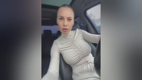 Media: Video of a young woman with light skin, straight hair pulled back, wearing a tight, light gray top with ruched sides, sitting in a car. Background shows blurred car interior and outdoor view.