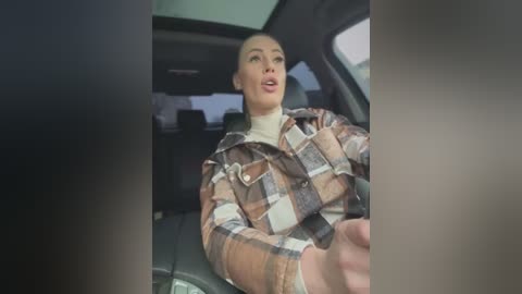 Video of a blonde woman with light skin, wearing a brown plaid coat, driving a car. Inside the car, the dashboard and window are visible, with a blurred background.